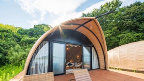 Una tienda de campaña abovedada con una terraza de madera, muebles de mimbre y un vidrio que da a un interior con una cama y una lámpara colgante.