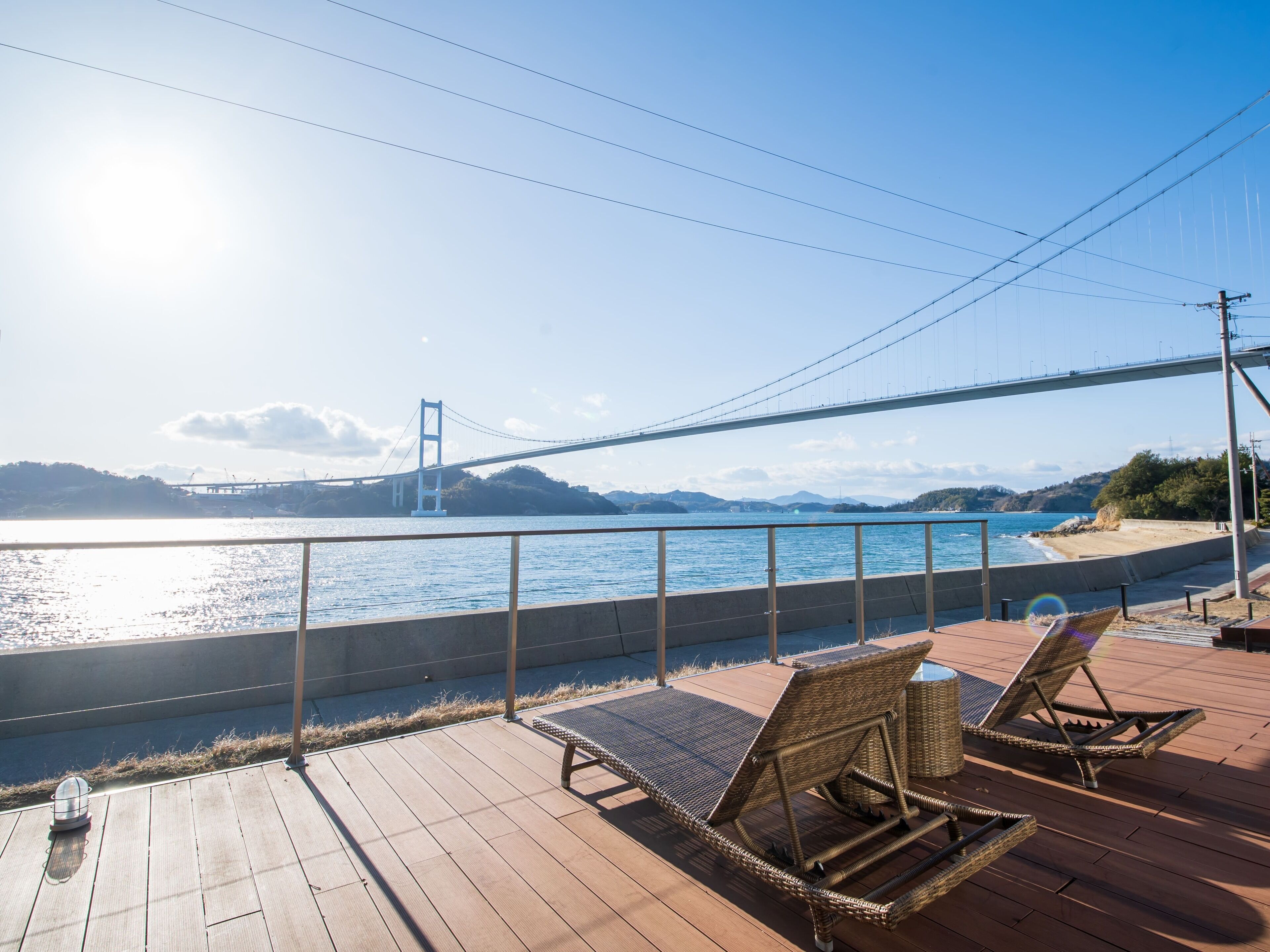 Una terraza de madera con sillones de mimbre que dan a un cuerpo de agua, un puente y colinas distantes.