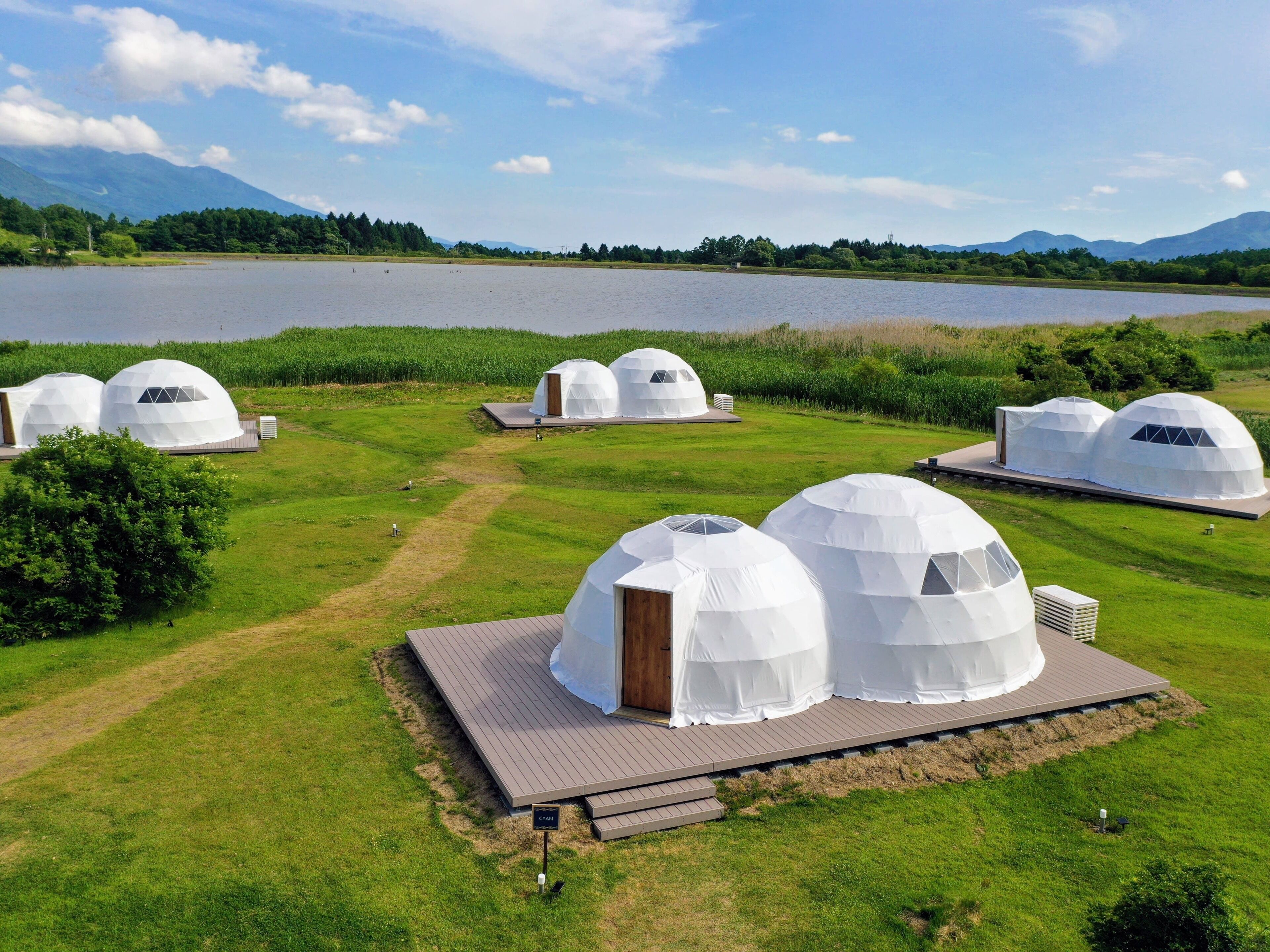A series of dome-shaped yurts with wooden decks, set in a grassy area near a lake with mountains in the background.