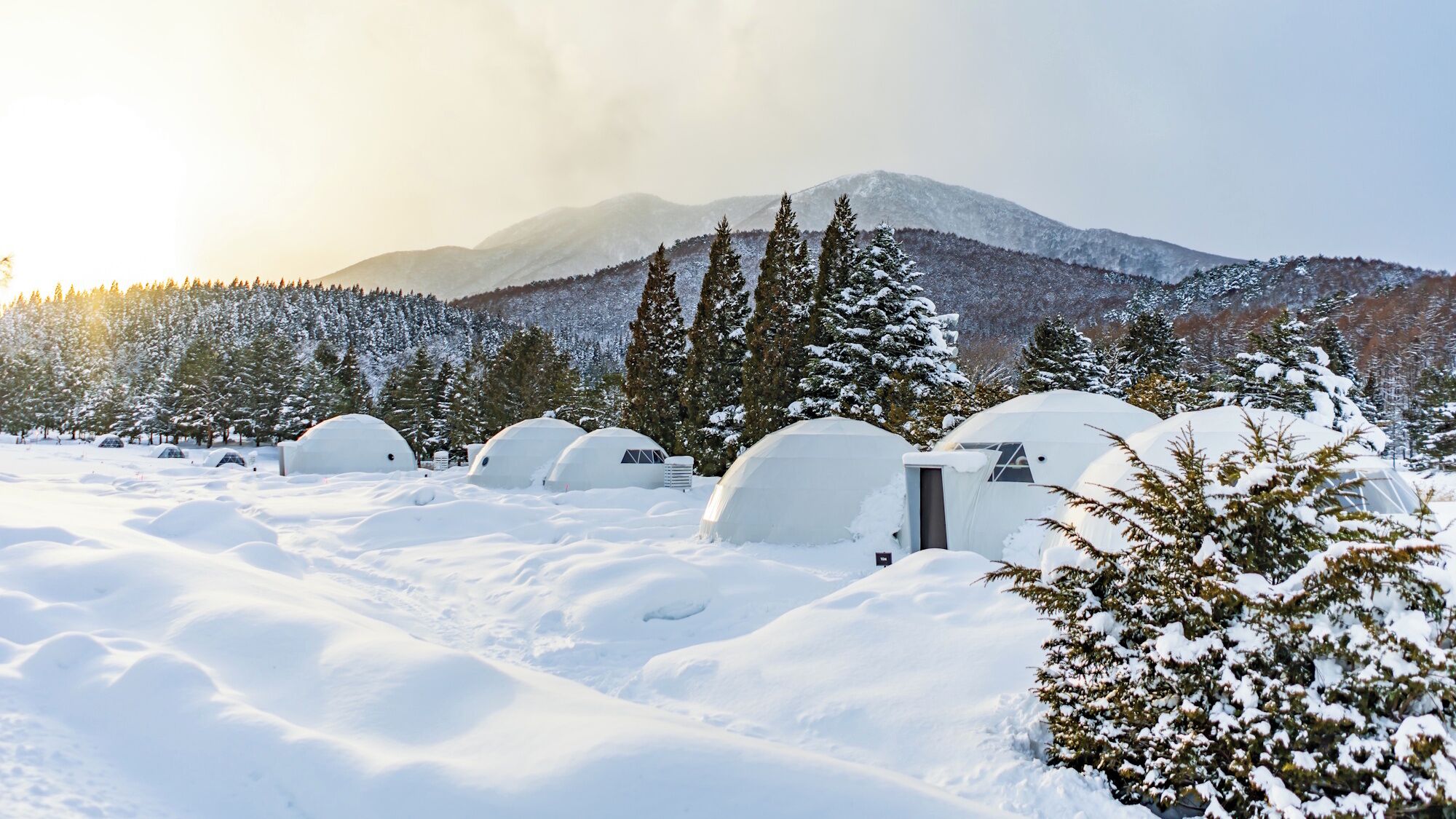 Schneelandschaft mit mehreren kuppelförmigen Bauwerken, vermutlich Iglus, vor verschneiten Bäumen und Bergen.