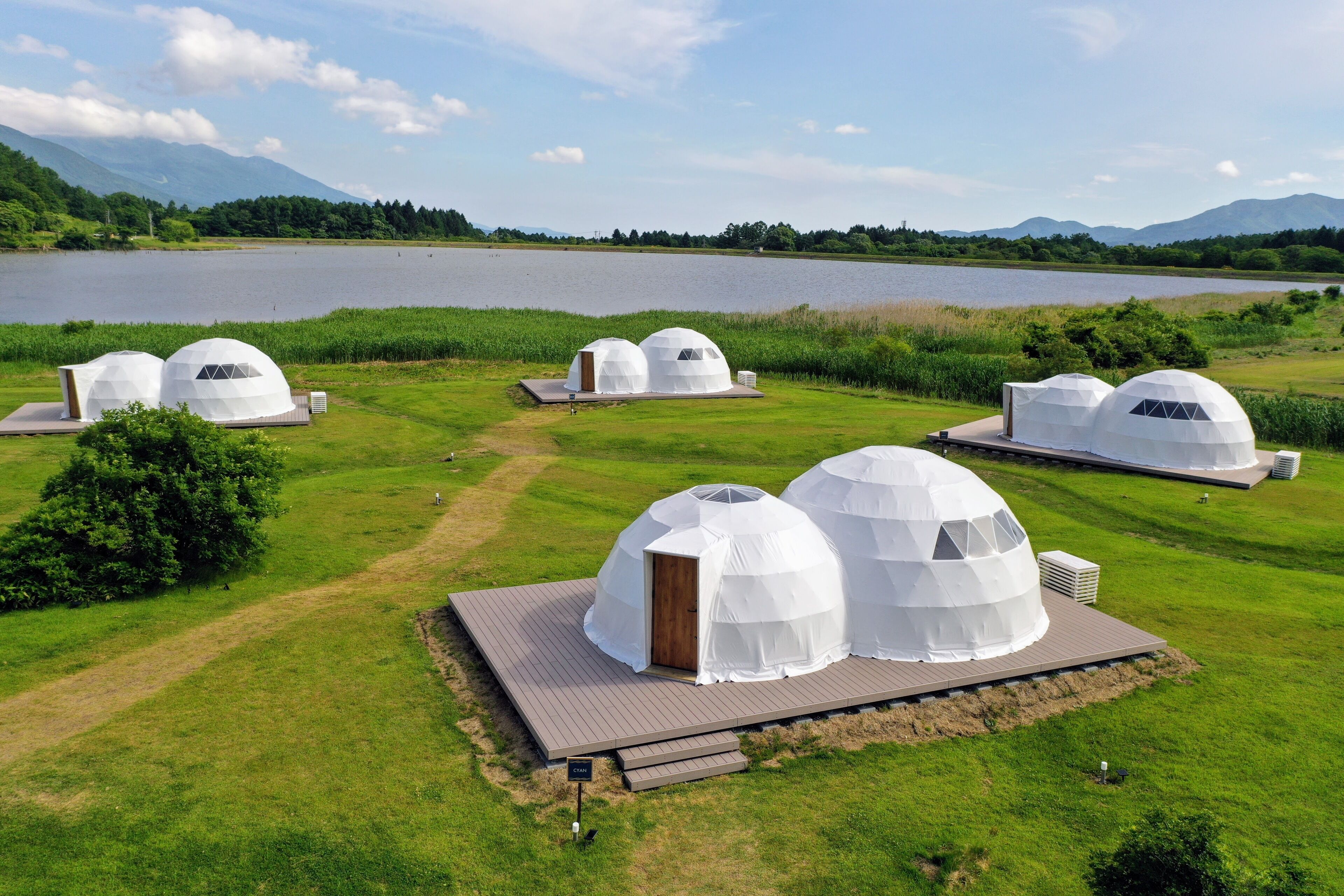 A series of dome-shaped yurts with wooden doors and air conditioning units, situated on a grassy area with a lake and mountains in the background.