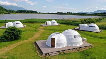 A series of dome-shaped yurts with wooden doors and air conditioning units, situated on a grassy area with a lake and mountains in the background.