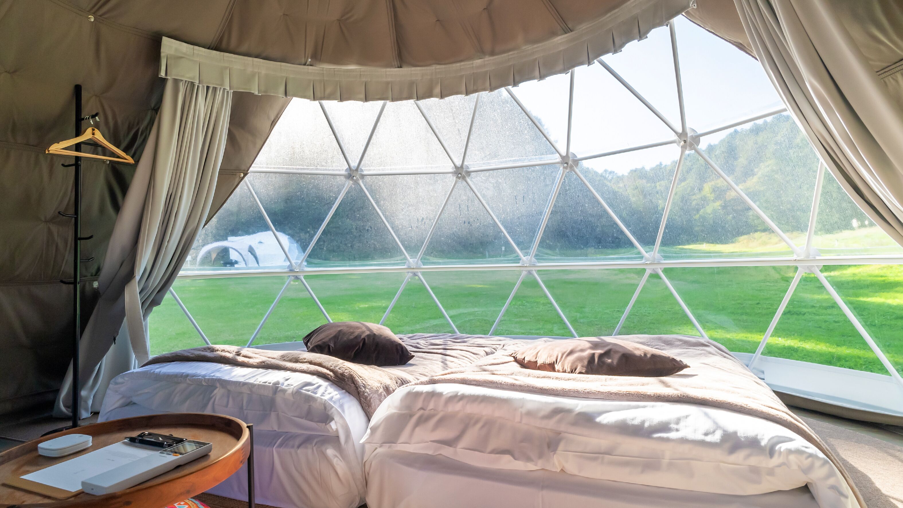A tent with a bed, a wooden hanger, a table with a book and a phone, and a view of a green field through a window.