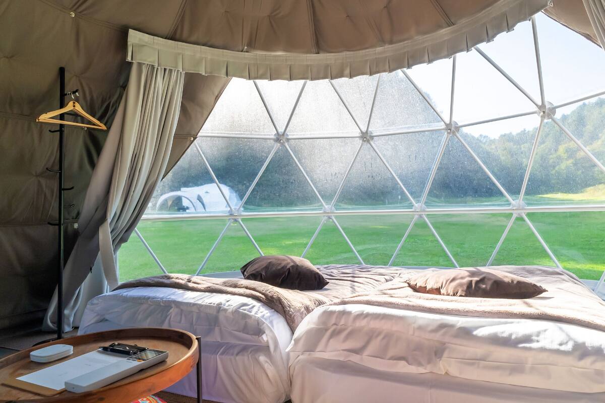 Ein Zelt mit Bett, einem hölzernen Kleiderbügel, einem Tisch mit Buch und Telefon sowie Blick auf ein grünes Feld durch ein Fenster.
