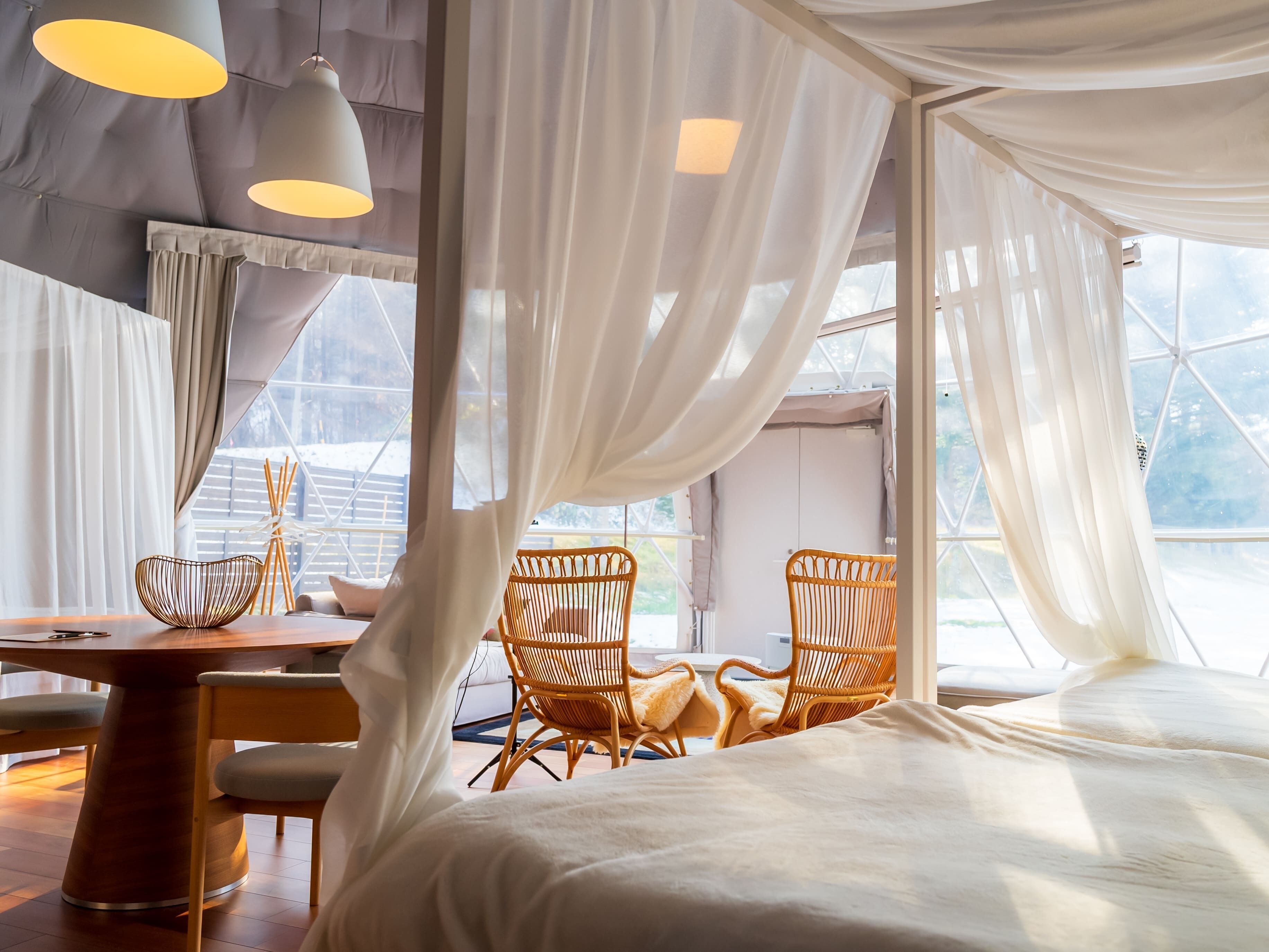 A room with a bed, two wicker chairs, a wooden table, and a large window with sheer curtains.
