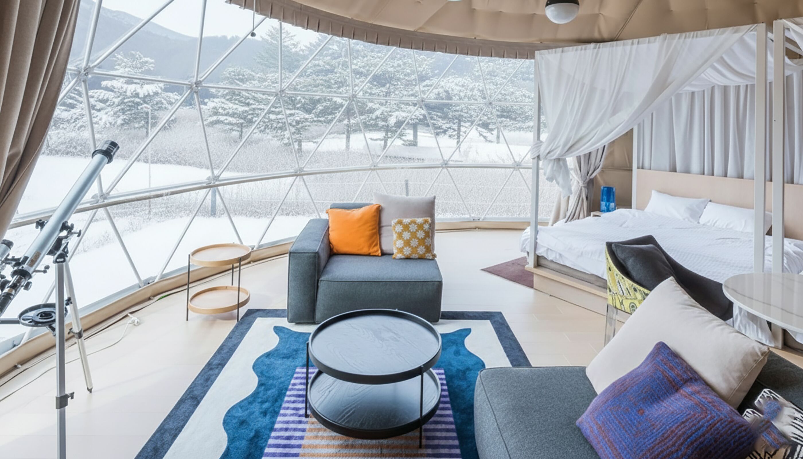 Une pièce moderne et bien éclairée, dotée d’une grande fenêtre offrant une vue sur un paysage enneigé. On y trouve un lit avec du linge de lit blanc, un canapé gris agrémenté de coussins colorés et une petite table ronde.