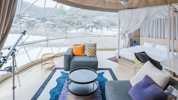 Une pièce moderne et bien éclairée, dotée d’une grande fenêtre offrant une vue sur un paysage enneigé. On y trouve un lit avec du linge de lit blanc, un canapé gris agrémenté de coussins colorés et une petite table ronde.