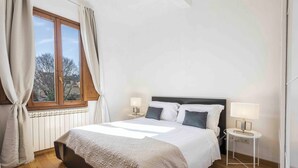 A bedroom with a large bed, two bedside tables with lamps, a window with curtains, and a radiator. - Casa Carolina by Mmega (Florence)