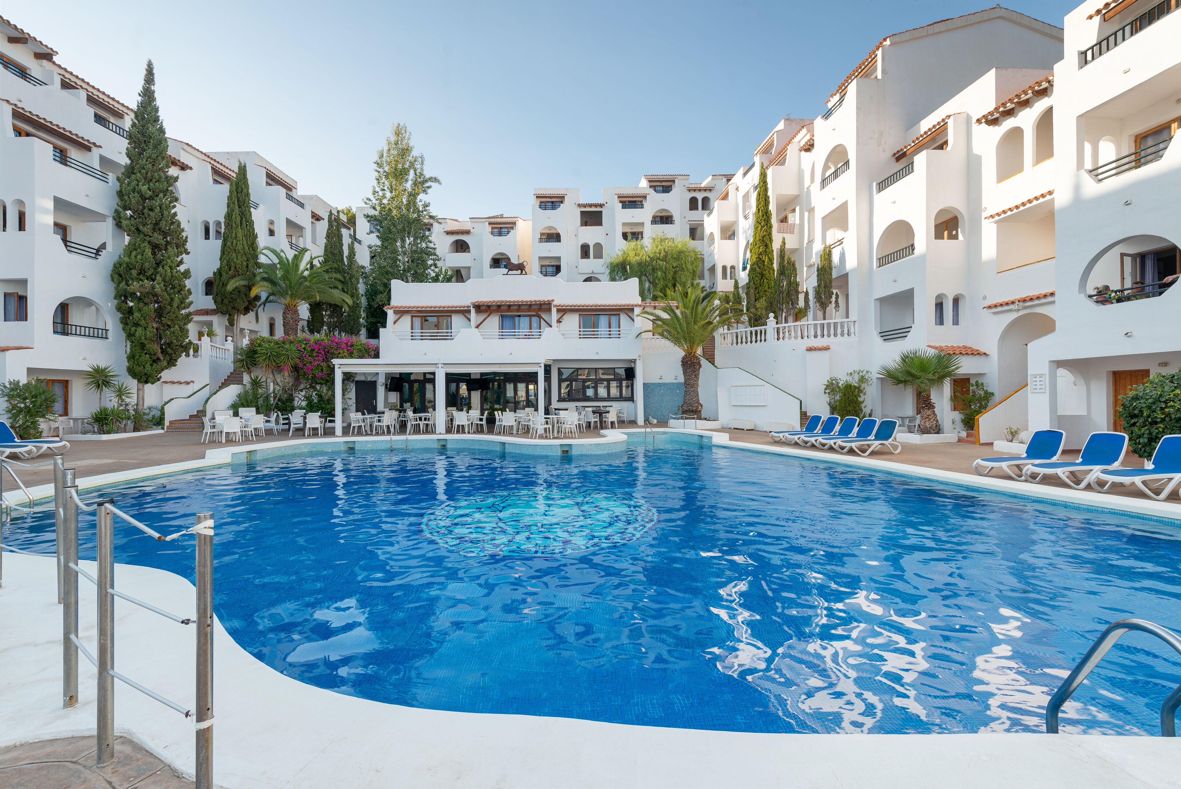 Una piscina circondata da edifici bianchi con balconi, palme e lettini.