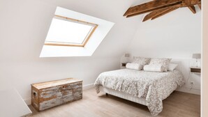 A bedroom with a slanted ceiling, a bed with a patterned bedspread, a wooden chest, and a small table with a lamp. - Welcome to Arsene Gardens (TINTÉNIAC)