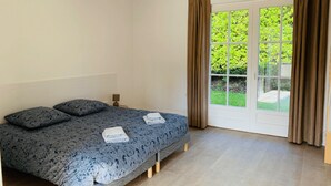 A bedroom with a bed, two pillows, a nightstand, and a window with curtains. - Welcome to Arsene Gardens (TINTÉNIAC)