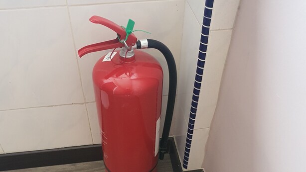 Extintor rojo montado en la pared en un rincón, con suelo de madera y paredes de azulejos.