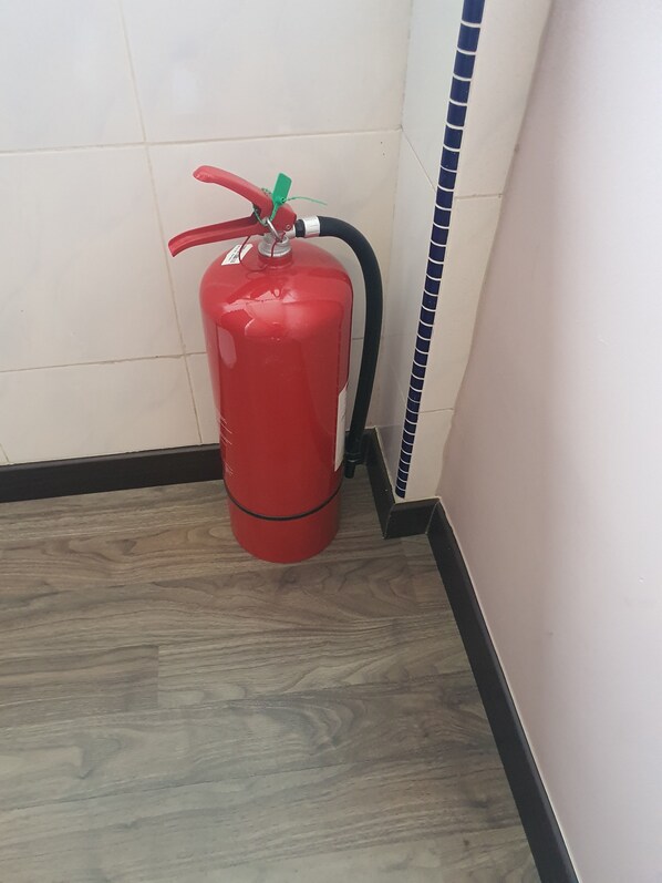 Extintor rojo montado en la pared en un rincón, con suelo de madera y paredes de azulejos.