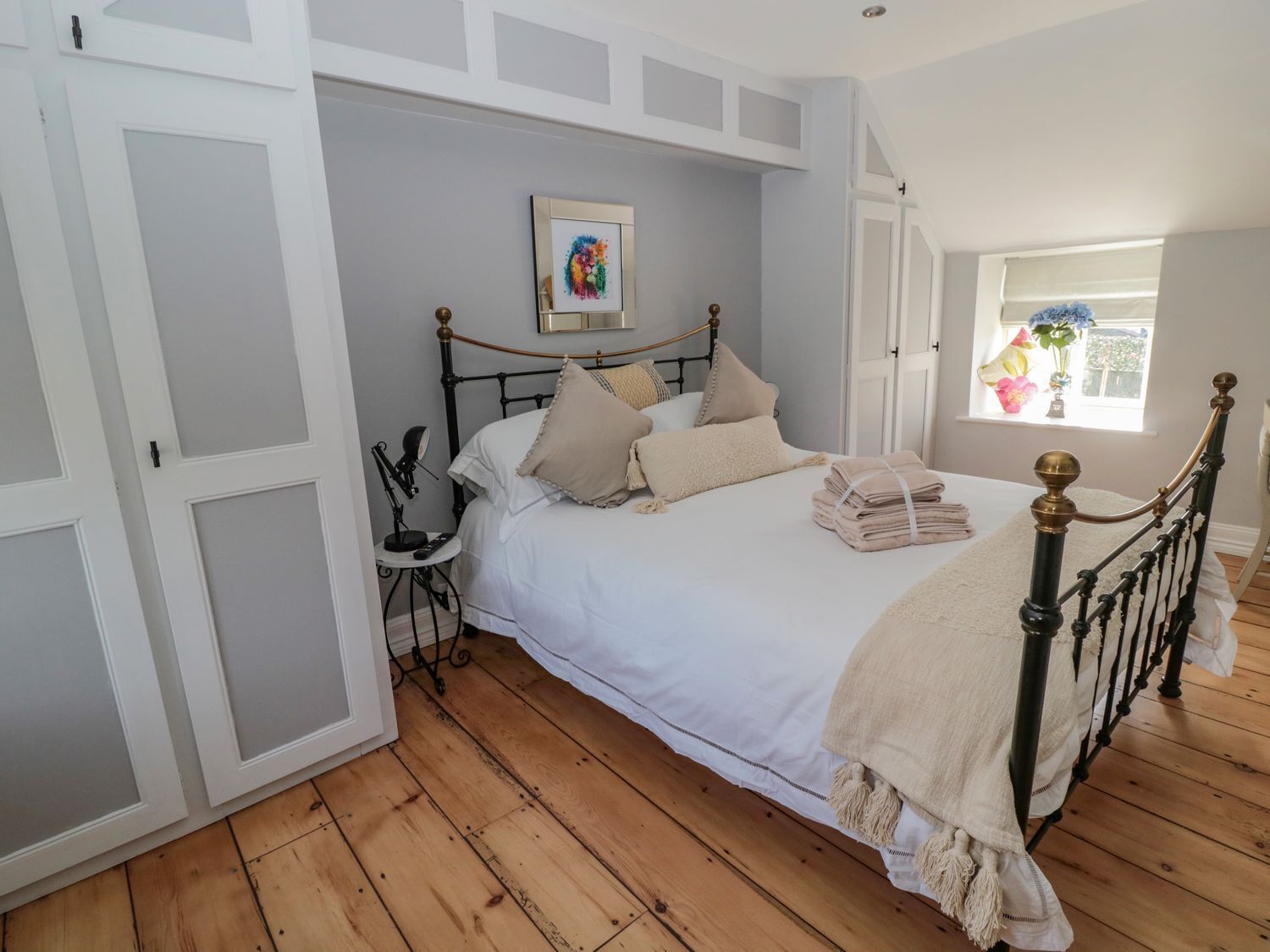 A bedroom with a bed, a bedside table, a lamp, a window with flowers, and a picture on the wall.