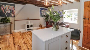 Une pièce avec un plancher en bois, une armoire blanche avec des tiroirs, un vase rempli de fleurs, un tableau représentant un cheval et un plafond en bois avec des poutres apparentes.