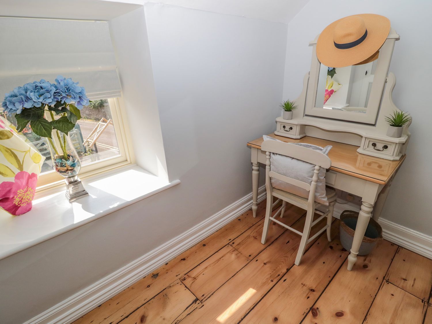 A room with a wooden desk, a chair, a vanity with a mirror, and a window with flowers.