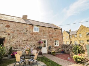 A stone building with a door and windows, a small garden with potted plants, and a table with chairs. - O'neil Cottage (Chathill)