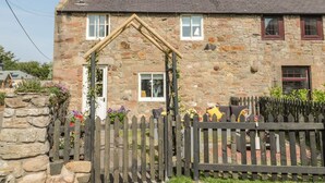 Une maison en pierre avec un toit à pignon, une clôture en bois et un jardin fleuri.