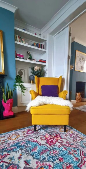 A room with a yellow armchair, a patterned rug, a bookshelf, and a fireplace.