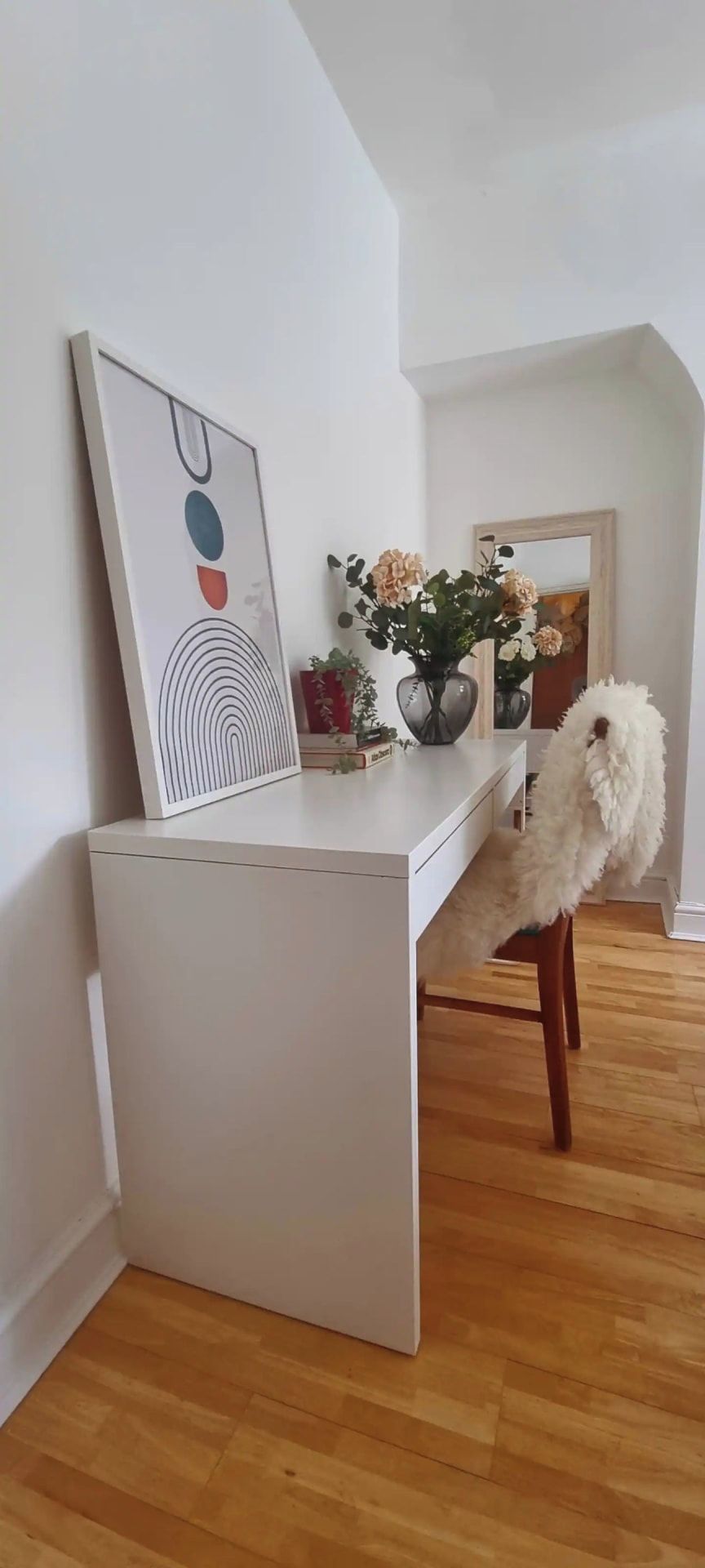 A white desk with a framed abstract painting, a vase with flowers, and a fluffy chair in a room with wooden flooring.
