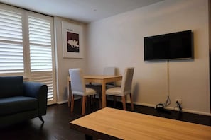 Uma sala de estar com sofá, mesa de jantar com cadeiras, televisão na parede e um quadro emoldurado.