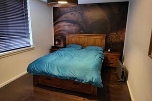 Quarto com cama de madeira, mesa de cabeceira, aquecedor fixado na parede e janela com persianas.