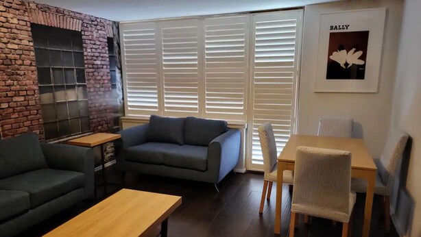 A living room with a brick wall, a grey sofa, a wooden coffee table, and a dining area with a chair and table. - Homely 1 Bedroom Retreat in South Yarra With Balcony (South Yarra)