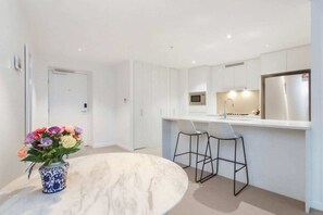 A modern kitchen with white cabinetry, a stainless steel refrigerator, a microwave, and a dining area with a marble table and two bar stools. - Luxury 1 Bedroom Retreat in Brisbane City With Pool and gym (Brisbane)