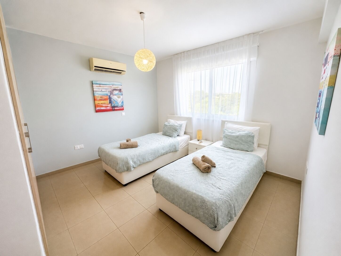 Un dormitorio con dos camas, ventilador de techo, aire acondicionado, un cuadro y una ventana con cortinas translúcidas.