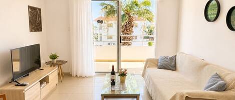 A modern living room with a flat-screen TV, a sofa, a coffee table, and a balcony with a view of palm trees.