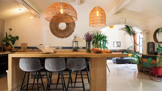 Een moderne keuken met een houten kookeiland, barstoelen, hangende rieten lampen en verschillende planten.