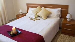 Una cama bien hecha con sábanas blancas, dos mesitas de noche con lámparas, una manta roja y una vela encendida sobre una bandeja.