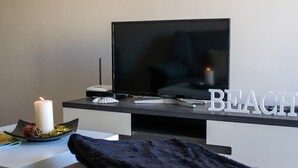 Ett modernt vardagsrum med en platt-TV, en vit dekorativ detalj som stavar ordet ”BEACH”, och en svart pläd på en soffa.