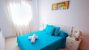 Un letto rifatto con una coperta e dei cuscini azzurri, un comodino bianco con una lampada e una bottiglia, e un quadro appeso al muro.