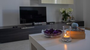 Un saló modern amb televisió de pantalla plana, un suport de vidre per a espelma amb espelma encès, i un bol amb flors morades sobre una taula blanca.