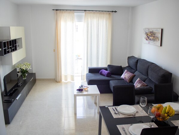 A modern living room with a grey sofa, a small coffee table, and a dining table set with plates and glasses. - PUESTA DE SOL RENTALS 3AR (PLAYA SAN JUAN)