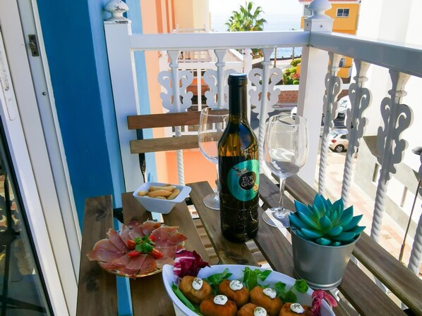 A balcony with a table set for dining, featuring a bottle of wine, glasses, a bowl of breadsticks, a plate of prosciutto, and a potted plant.