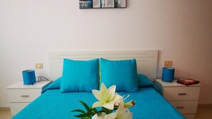 Un lit bien fait, recouvert d’une couvre-lit bleu vif et agrémenté d’oreillers assortis, une table de chevet blanche avec une lampe, et un joli arrangement floral blanc posé sur le lit.