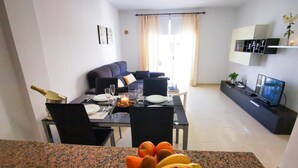 Un salon moderne comprenant un coin repas, un canapé, un téléviseur et un panier de fruits posé sur la table.