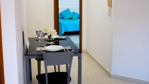 A narrow hallway with a dining table set for two, a chair, and a vase of white flowers. A blue sofa is visible in the background, and a telephone is mounted on the wall. - PUESTA DE SOL RENTALS 2BR (PLAYA SAN JUAN)