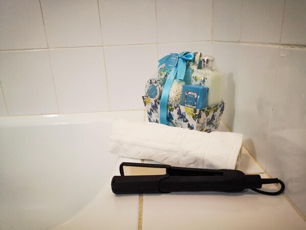A bathroom with a white bathtub, a floral-patterned gift basket with a blue ribbon, a white towel, and a black hairdryer. - PUESTA DE SOL RENTALS 2AR (PLAYA SAN JUAN)