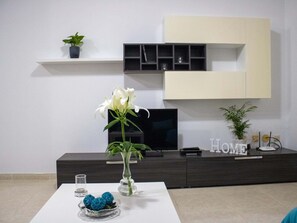 A modern living room with a white table, a vase with a white flower, a decorative blue object, a dark wooden cabinet, and a wall-mounted TV.