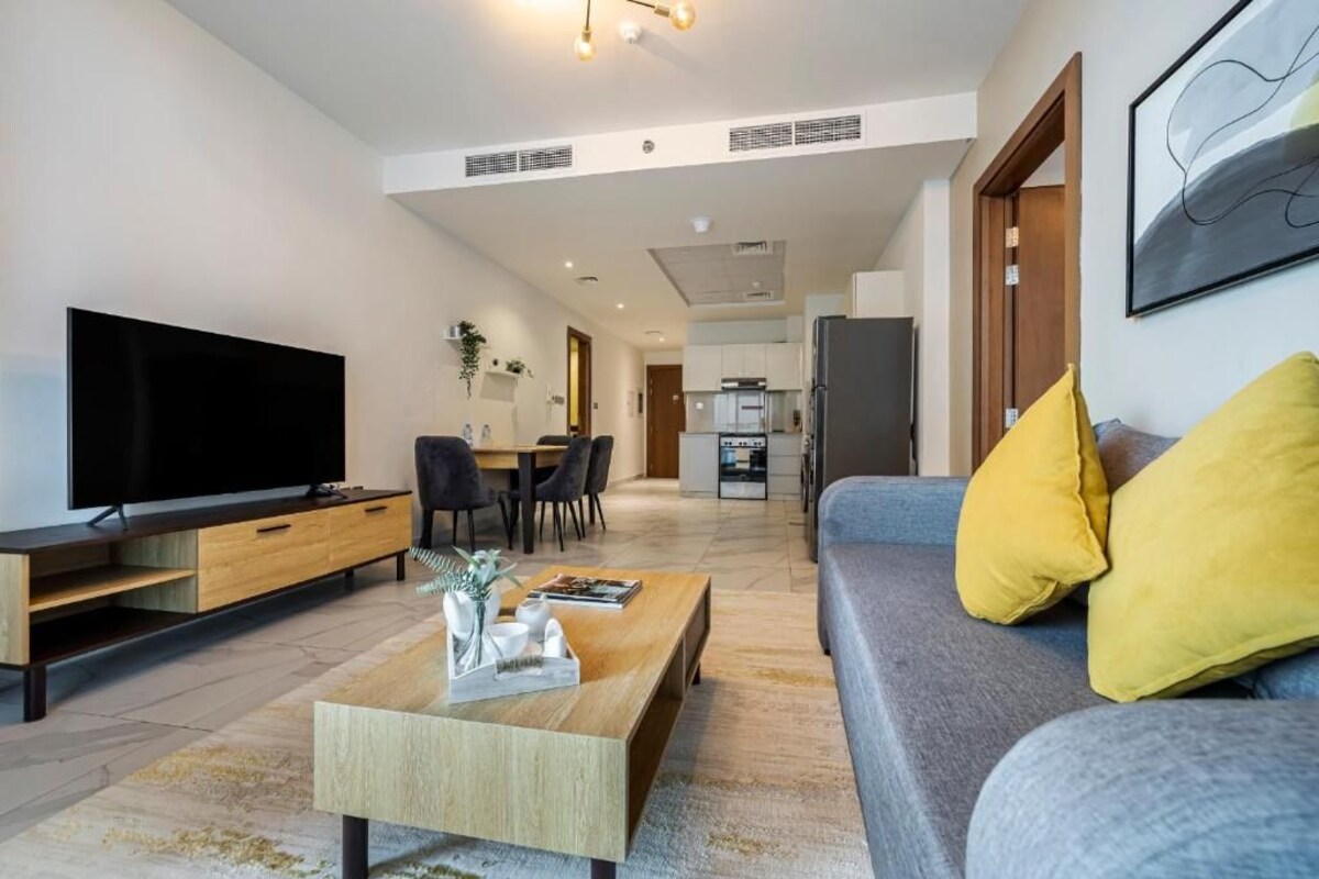 Una sala de estar moderna con una televisión de pantalla plana, una mesa de centro de madera, un sofá gris con cojines amarillos y un área de comedor al fondo.