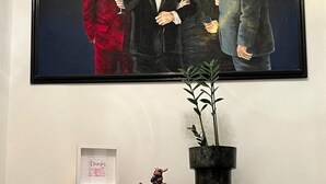Un tableau encadré représentant quatre hommes en costume, une console en bois avec des tiroirs, une plante en pot et un chien allongé sur un tapis.