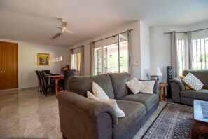 Ein geräumiges Wohnzimmer mit einer Sofagarnitur, einem Esstisch mit Stühlen, einem Deckenventilator und großen Fenstern mit Vorhängen.