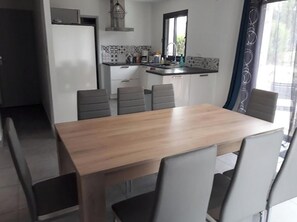 Uma área de refeições com mesa de madeira e seis cadeiras, uma cozinha com geladeira e pia, e uma janela com cortinas.