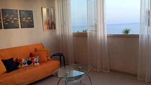 A living room with an orange sofa, a glass table, and a view of the sea through sheer curtains. - A terrace overlooking the sea 19084041C226886 (Sicilia)
