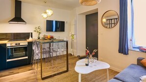 A modern kitchen with a central island, a dining area with clear chairs, a small white table with a bottle and glasses, and a living area with a sofa and a round mirror on the wall.