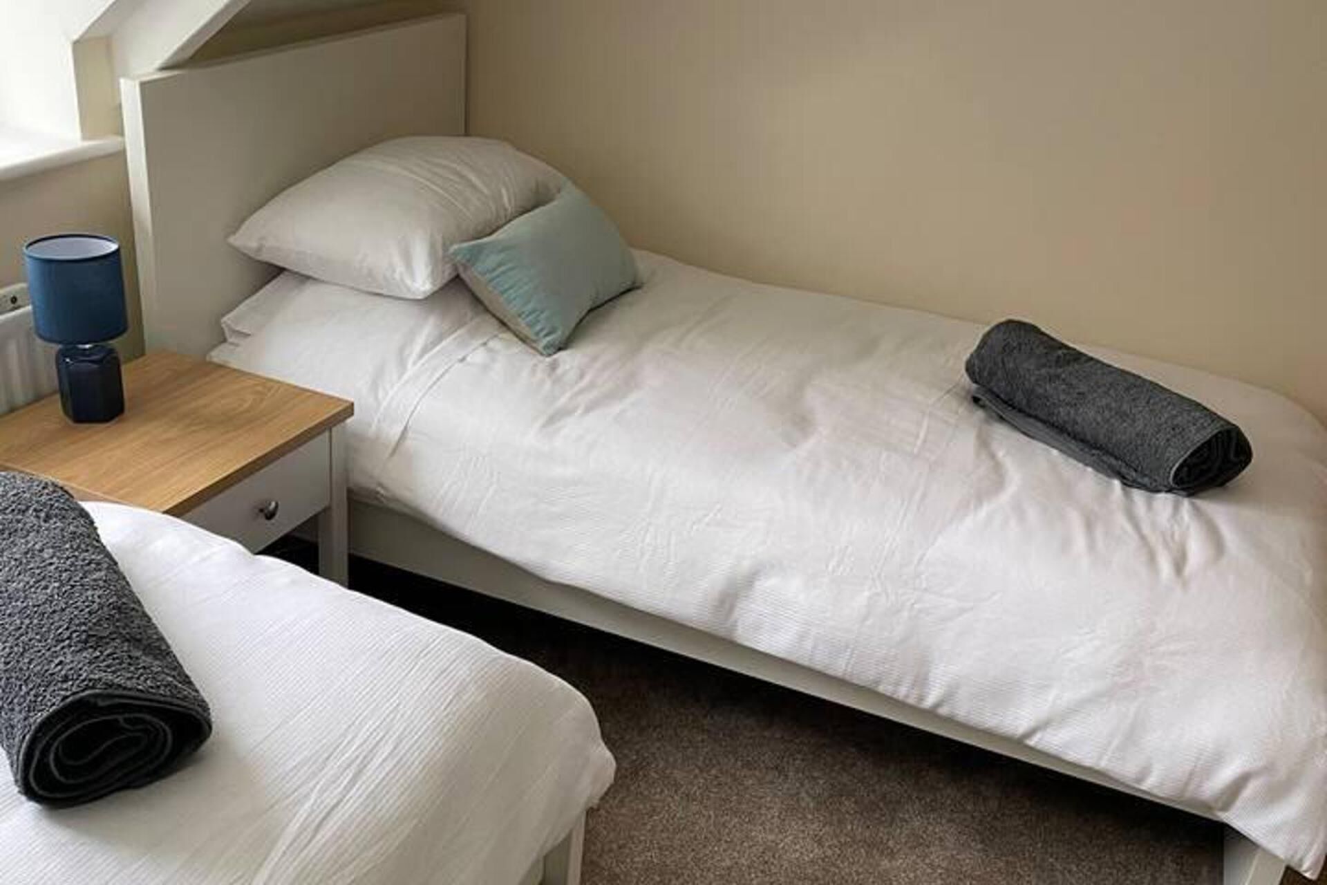 A single bed with white bedding, a blue pillow, and a rolled-up grey towel on the bed.