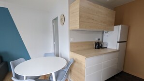 A compact kitchen with a white countertop, wooden cabinets, and a white refrigerator. There is a round table with two chairs and a clock on the wall. - Nice little house close to the sea and town center (La Rochelle)